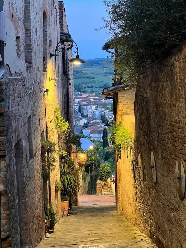 Narrow cobblestone street in an Italian village at sunset with warm lights and a view over the valley