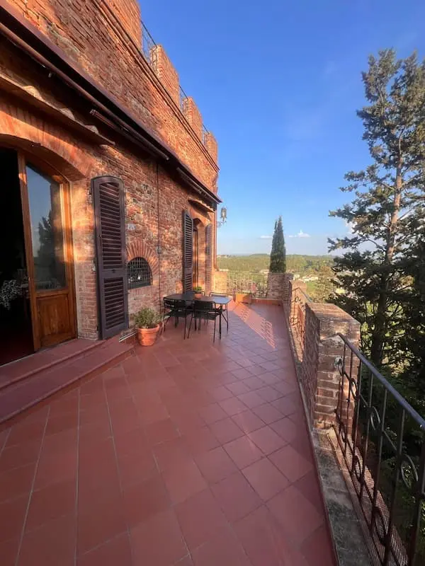 Terrace of a Tuscan house with outdoor table, brick walls and open views over hills and countryside.