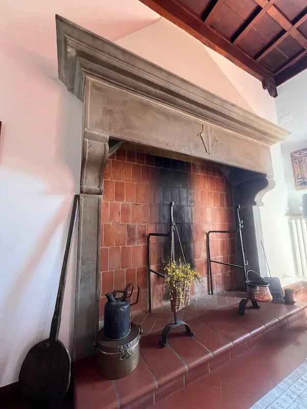 A monumental hand-carved stone fireplace with a red brick interior and antique kitchen utensils in a historic Tuscan estate in Certaldo Alto.